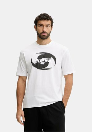 Uomo con la barba che indossa una maglietta bianca a maniche corte con il logo "GUESS" a vortice in bianco e nero, in piedi con una mano in tasca su uno sfondo semplice.