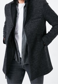 Abrigo a cuadros gris y negro, con cuello con muesca, cierre de un solo botón, bolsillos frontales y tela texturizada. Superpuesto sobre una camisa gris.