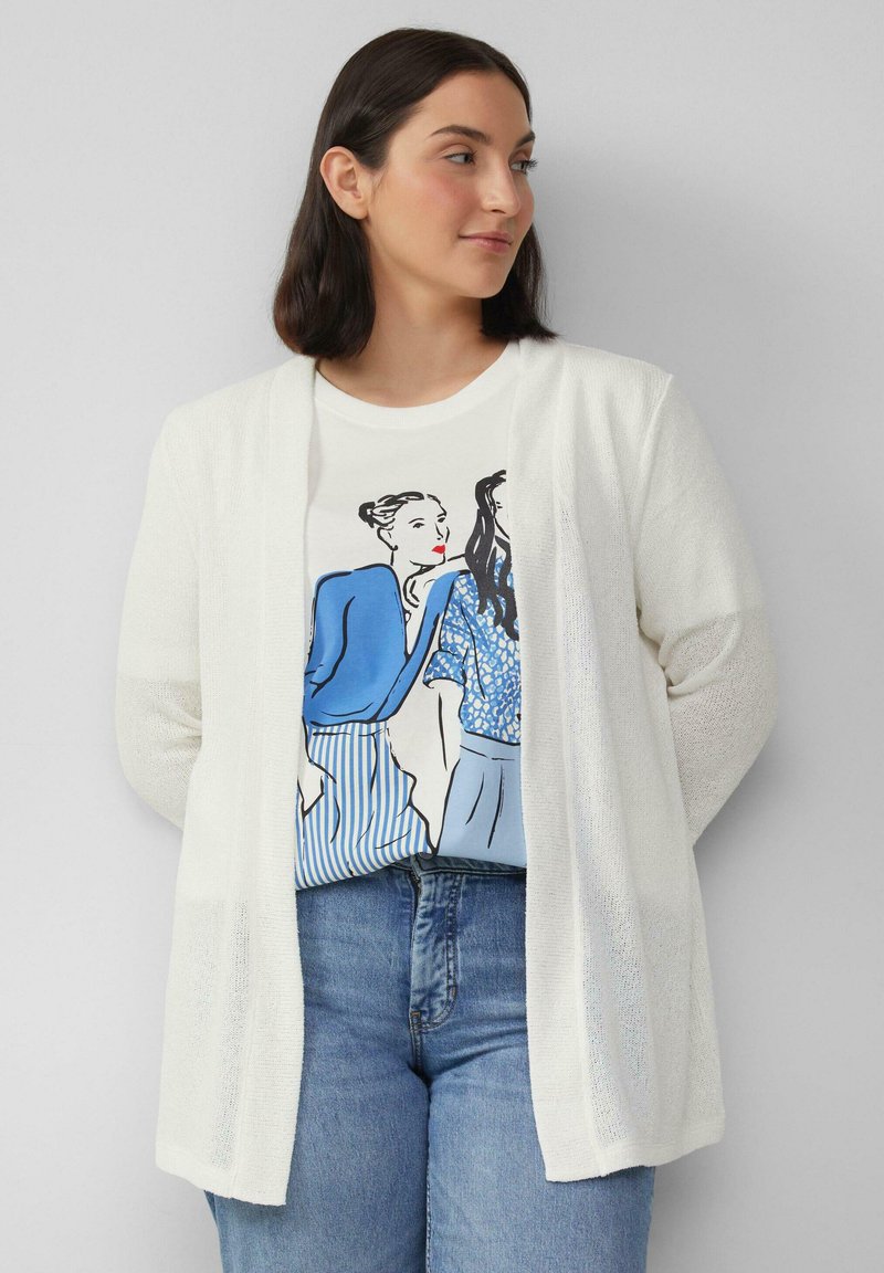 Leichte, offene Strickjacke in Weiß mit gestrickter Textur, kombiniert mit einem blauen Grafik-T-Shirt mit illustrierten Designs. Klassische blaue Jeans runden das Outfit ab.