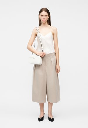 Kvinne iført hvit camisole, beige vide, avkortede bukser, svarte spisse sko og bærende på hvit skulderveske med dekorativ stropp.