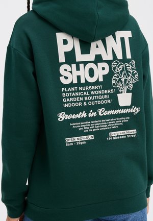 Felpa verde con testo e grafica bianca. Il design sul retro include "PLANT SHOP", illustrazioni di piante e dettagli aziendali. Materiale in cotone.