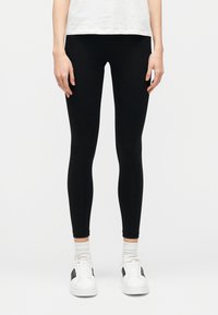 Leggings nere con una texture liscia ed elastica, design aderente e vita alta, abbinati a sneaker bianche e calzini sportivi bianchi.