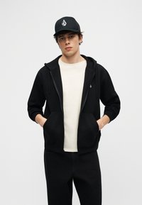 Jeune homme portant une casquette de baseball noire, un sweat à capuche noir avec fermeture éclair sur un T-shirt blanc, les mains dans les poches, debout devant un fond gris uni.