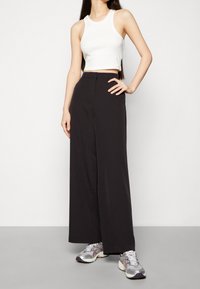 Kvinna klädd i en vit ärmlös crop top, högmidjade vidbyxor med vida ben i svart och metallicsöta silverfärgade löparskor, som står med ena handen på höften.