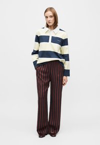 Polo cu mânecă lungă cu dungi albastre închis și galben deschis; are guler alb și logo. Asortat cu pantaloni largi cu dungi bordo și negre.