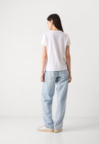 T-shirt blanche à manches courtes avec un col rond, associé à un jean bleu clair à coupe ample, et des baskets beiges, vu de dos.