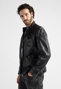 Zwarte leren jasje met een ritssluiting aan de voorkant, zijzakken en gestructureerde schouderdetails. Glad afgewerkt en strak ontwerp, gecombineerd met grijze jeans.