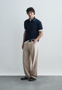 Μπλε ναυτικό πόλο μπλουζάκι με λευκές λεπτομέρειες, μπεζ παντελόνι φαρδύ και μαύρα παπούτσια. Το μοντέλο στέκεται με το ένα χέρι στην τσέπη, μπροστά σε ανοιχτό φόντο.