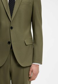 Chaqueta de traje verde oliva con textura suave, cierre de un solo botón, sutil bolsillo en el pecho y botones negros; combinada con pantalones a juego.