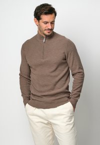 Brauner Strickpullover mit einem Reißverschluss und Mock-Neck, eng anliegenden Ärmeln und strukturiertem Muster. Kombiniert mit hellen Hosen.