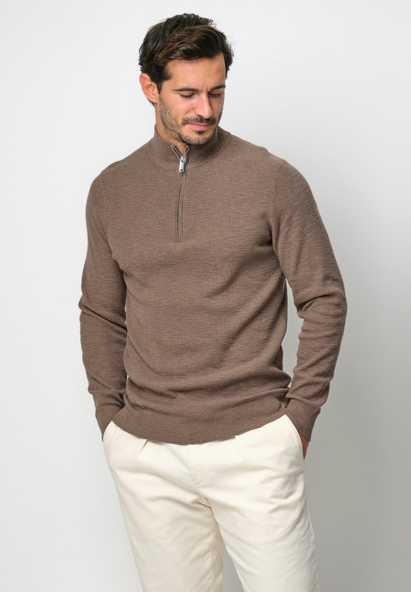 Brauner Strickpullover mit einem Reißverschluss und Mock-Neck, eng anliegenden Ärmeln und strukturiertem Muster. Kombiniert mit hellen Hosen.