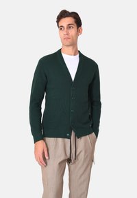 Cardigan verde scuro lavorato a maglia con scollo a V, cinque bottoni e texture a coste, indossato sopra una t-shirt bianca e pantaloni chiari.