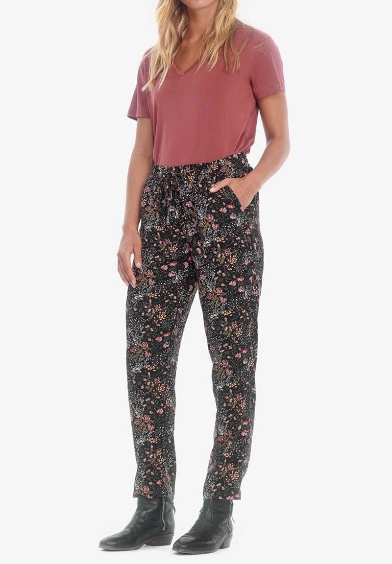 Donna che indossa un top a maniche corte color ruggine, pantaloni floreali neri a vita alta con cintura a fiocco e stivali neri alla caviglia, in piedi con una mano in tasca.