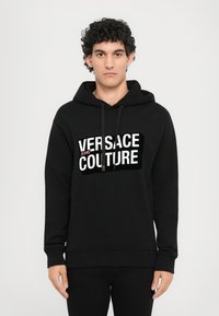 Čierna mikina z materiálu na mikiny s bielym grafickým logom "VERSACE JEANS COUTURE" na prednej strane. Klasický dizajn s kapucňou na sťahovanie.