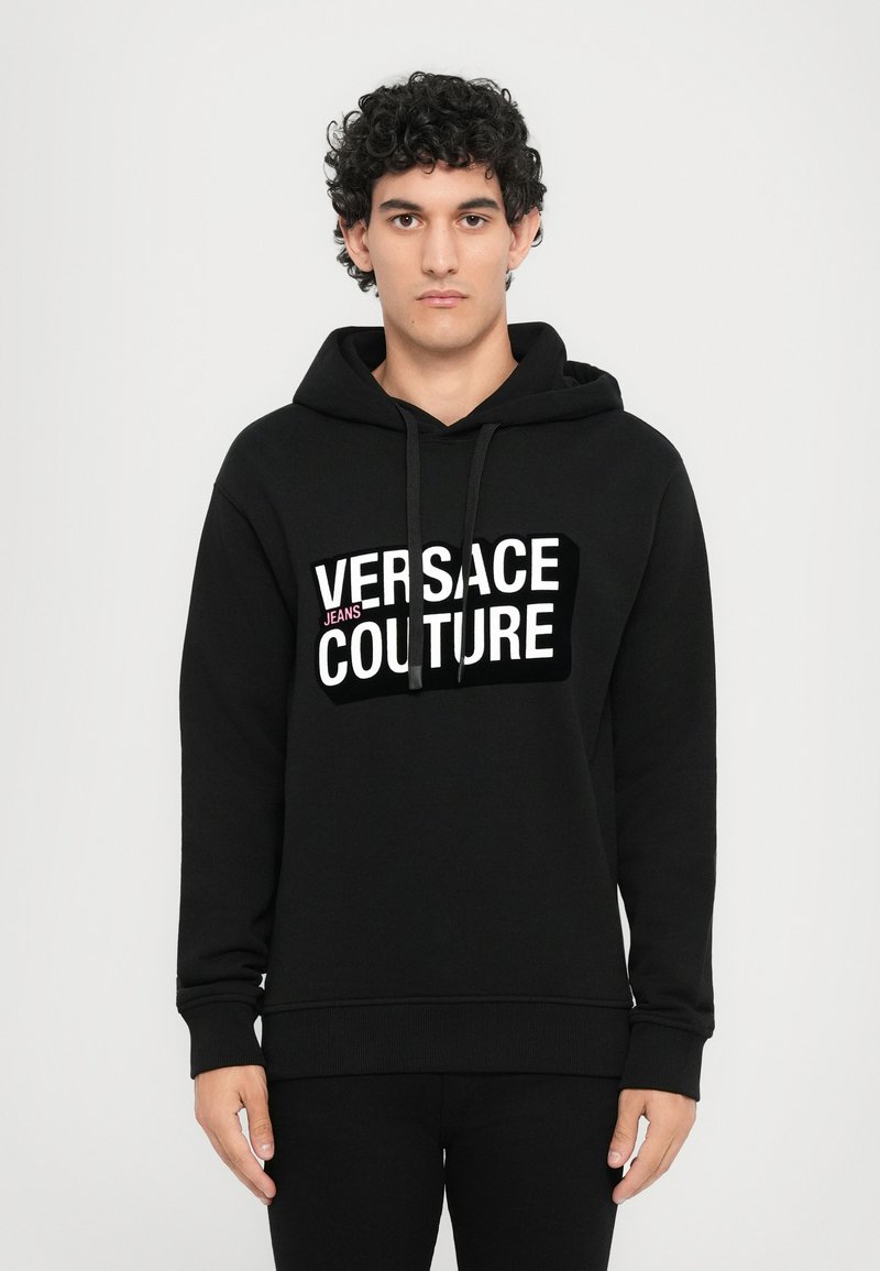 Čierna mikina z materiálu na mikiny s bielym grafickým logom "VERSACE JEANS COUTURE" na prednej strane. Klasický dizajn s kapucňou na sťahovanie.