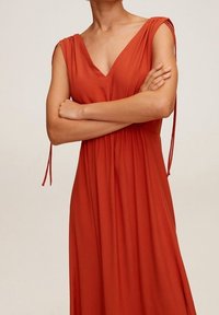 Femme en robe sans manches couleur rouille avec des bretelles nouées, bras croisés, sur un fond clair uni.