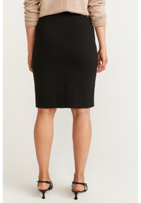 Černá tužková sukně s přiléhavým střihem, hladkou texturou a rozparkem vzadu, doplněná černými sandály na tenkých podpatcích. Neutrální svetr částečně viditelný.