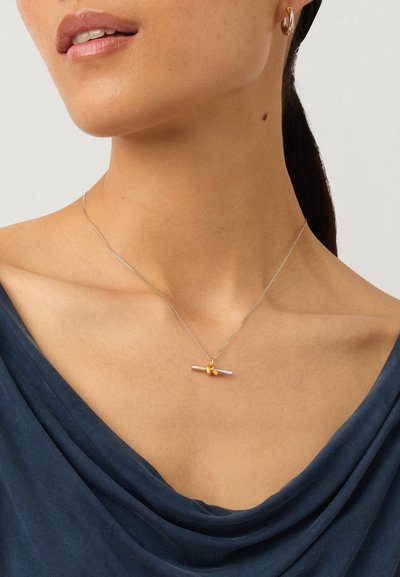 Gros plan d'une femme portant un collier en argent avec une barre horizontale et trois petits charms dorés, associé à une boucle d’oreille créole en or et un haut bleu marine.