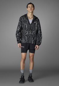 Leichte schwarze Windjacke mit einem strukturierten Camouflage-Muster, kombiniert mit schwarzen Shorts und gestreiften Socken. Sportliche Schuhe runden den Look ab.