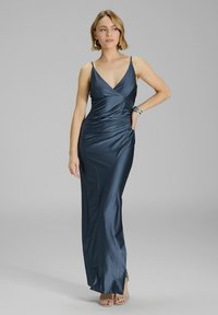 Robe maxi en satin navy avec décolleté en V et fines bretelles, présentant des drapés sur le côté et une fente haute sur la cuisse, associée à des talons à lanières.