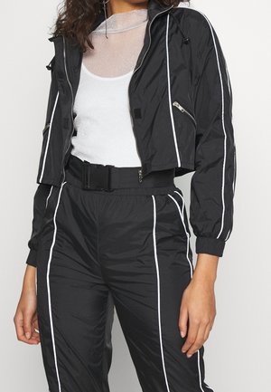Personne portant une veste courte noire et un pantalon avec un passepoil blanc, une taille blanche ceinturée et un top blanc transparent en dessous.