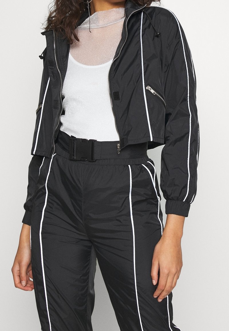 Personne portant une veste courte noire et un pantalon avec un passepoil blanc, une taille blanche ceinturée et un top blanc transparent en dessous.