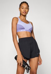 Lilla sports-bh med rund kutting og Nike-logo, sammen med svarte elastiske shorts med sideåpninger og Nike-emblem. Svart drikkeflaske i hånden.