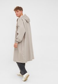 Abrigo impermeable beige con capucha, de mangas largas, puños elásticos y corte relajado, combinado con jeans oscuros y zapatillas blancas.