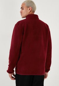 Maglione in pile bordeaux con colletto alto, maniche lunghe, texture liscia e vestibilità rilassata, progettato per calore e comfort.