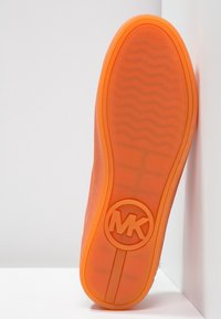 Oranžová gumová podrážka s texturovaným vzorem a kruhovým logem "MK" na spodní straně. Hladký povrch s zaobleným okrajem a mírnou flexibilitou.