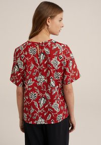 Blusa rossa con motivo floreale in bianco e nero, dotata di maniche corte e dettaglio a chiave sul retro. Tessuto dalla texture liscia.
