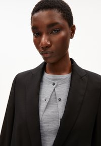 Vrouw met kort, krullend haar die een zwarte blazer over een lichtgrijs blouse-top draagt, recht vooruitkijkend met een neutrale uitdrukking tegen een witte achtergrond.