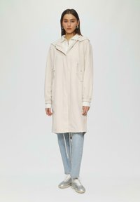Jasno-beżowy długi płaszcz z kapturem, z zamkiem błyskawicznym z przodu, dwiema bocznymi kieszeniami i elastycznymi mankietami, noszony na białej bluzce, dopasowany do niebieskich dżinsów.