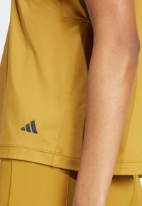 Camiseta deportiva de color mostaza con mangas cortas, que presenta una tela texturizada y un logotipo oscuro en la parte inferior izquierda.