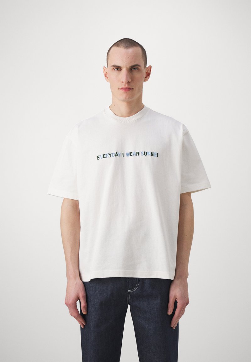 T-shirt en coton blanc avec des manches courtes, un col rond et un texte brodé "TOUS LES JOURS JE PORTE SUNNE" en bleu et vert.
