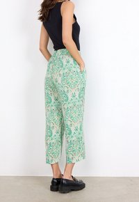 Pantalones de pierna ancha con estampado de paisley en verde y cintura alta, que cuentan con bolsillos y una tela ligera. Combinados con un top sin mangas negro y zapatos negros.