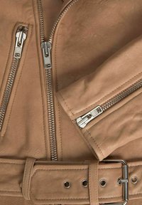 Tan spalvos odinė striukė su sidabriniais užtrauktukais ir atitinkančia diržu. Dizainas pasižymi lygiu paviršiumi, elegantiškais kontūrais ir matomais siuvimo detalėmis.