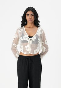 Witte bloemenkant crop top met een V-hals, trekkoord aan de voorkant en lange mouwen, gecombineerd met zwart, structuur wide-leg broek.
