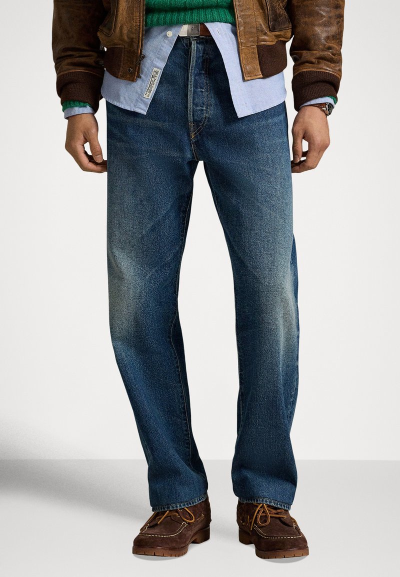 Μπλε τζιν με άνετη γραμμή, ελαφρύ ξεθώριασμα και �σχεδιασμό με πέντε τσέπες, φορεμένο με καφέ δερμάτινο μπουφάν και καφέ παπούτσια.