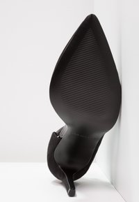 Scarpa nera con tacco alto, punta affilata e suola nera texturizzata, con una parte superiore in morbido camoscio e una silhouette elegante.