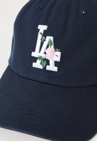 Navy cap featuring embroidered "LA" letters with floral details in pink and green. The cap has a curved brim and textured fabric.