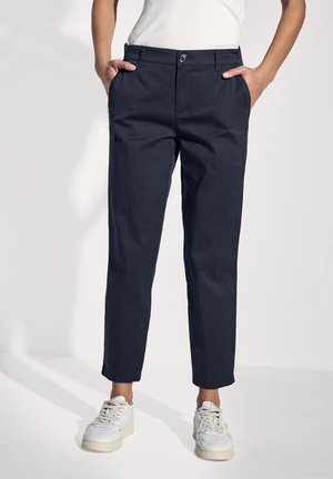 Vrouw draagt donkerblauwe rechte broek, witte sneakers en een wit topje, staand met handen in de zakken tegen een effen achtergrond.