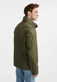 Oliwkowa kurtka nylonowa z wysokim kołnierzem, bocznymi kieszeniami i luźnym krojem, zestawiona z jasnoniebieskimi dżinsami.