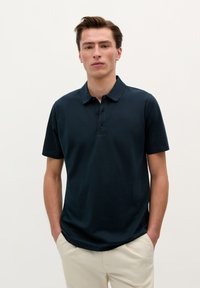 Námorně modré polo tričko vyrobené z m�ěkké bavlny, se zapínáním na tři knoflíky, krátkými rukávy a volným střihem. Kombinováno se světlými kalhotami.