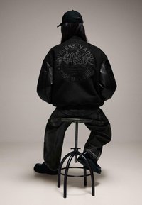 Fekete, túlméretezett bomberdzseki bőr ujjakkal és dombornyomott hátrész dizájnnal. A modell sötét farmernadrágot és fekete csizmát visel.