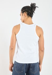 White, ribbed tank top with wide shoulder straps. The fabric appears soft with a snug fit, contrasted by dark denim jeans.