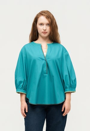 Moteris su ilgais rusvai raudonais plaukais, dėvinti laisvą turkio spalvos palaidinę su 3/4 pūstomis rankovėmis ir tamsiai mėlynas džinsus, stovinti ant vientisos fono.