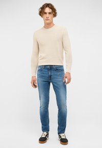 Beżowy sweter z prążkowanej dzianiny, wąskie niebieskie dżinsy, czarnoniebieskie sneakersy. Prosty design, miękka tekstura, casualowy krój z widocznymi detalami szycia.