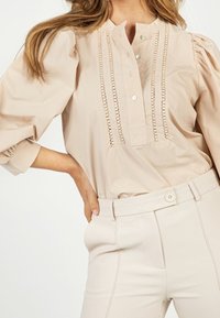 Blouse en coton beige clair à manches bouffantes, patte de boutonnage et détails de couture décorative. Associée à un pantalon taille haute assorti.