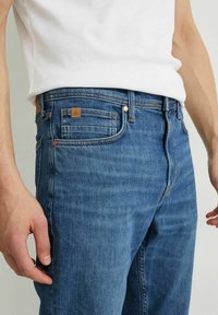 Pantalones vaqueros de mezclilla azul con corte recto, que cuentan con remaches de cobre, un parche de cuero en la parte trasera y un diseño clásico de cinco bolsillos.